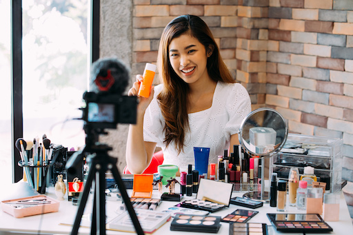 View of an influencer’s phone camera and lighting rig as they demonstrate beauty products in the background for social media.